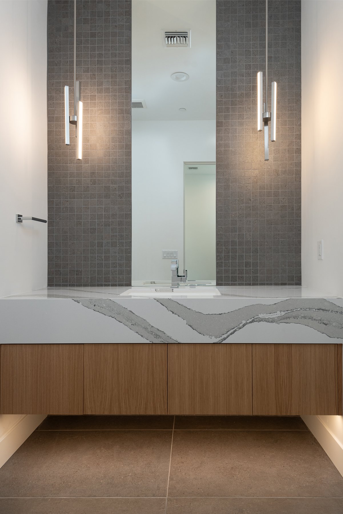 Modern bathroom vanity with a large rectangular mirror, gray tile backsplash, wooden cabinetry, and integrated sink. Two minimalist pendant lights hang beside the mirror.