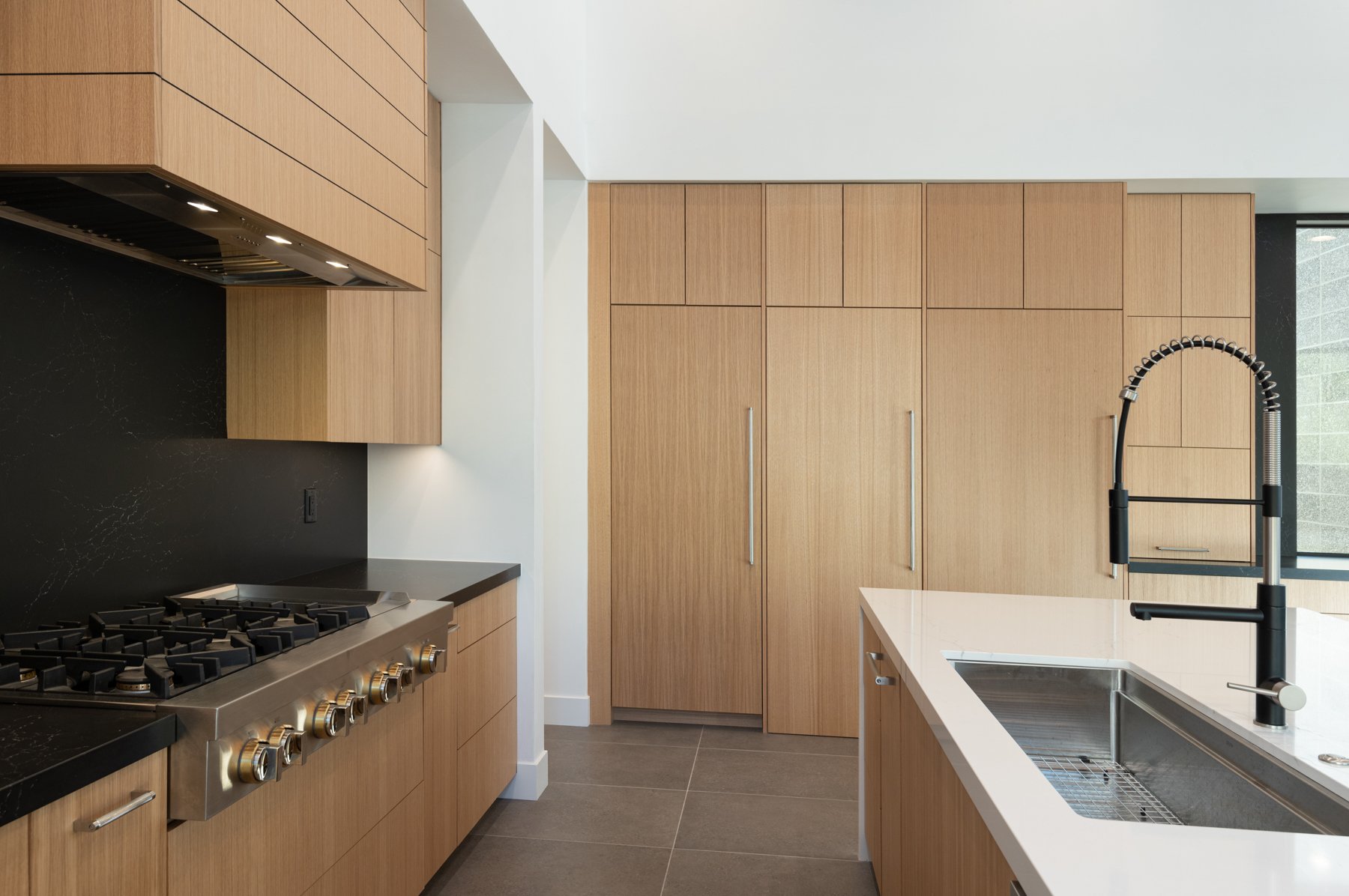 Modern kitchen with light wood cabinetry, large gas stovetop, black backsplash, and an island with a stainless steel sink and industrial-style faucet.