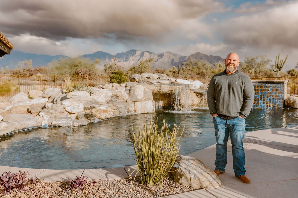 A man stands by a pool with a waterfall feature, surrounded by rocks and desert plants, under a cloudy sky with mountains in the background.