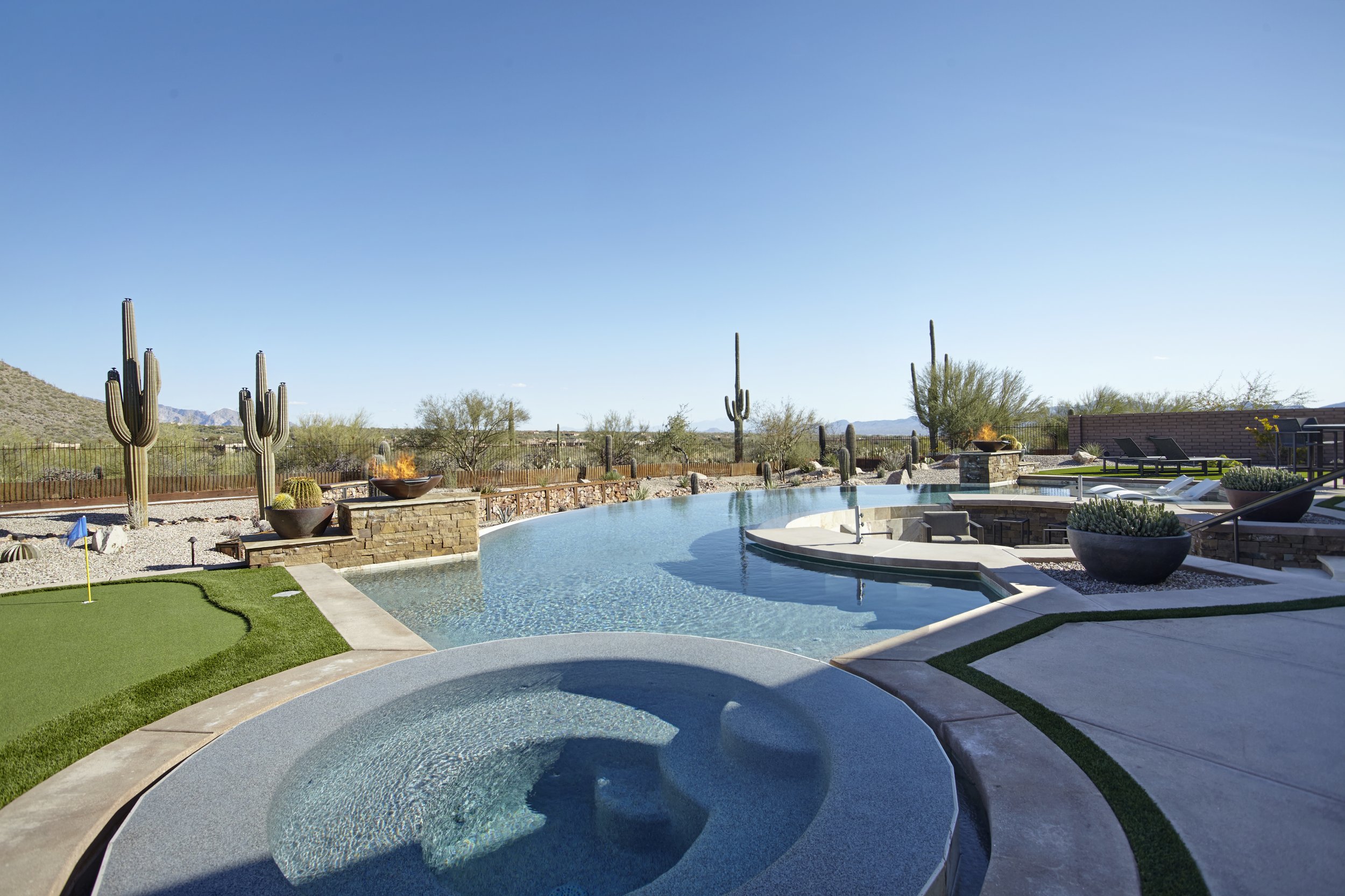 Luxury backyard with a swimming pool, spa, and desert landscaping featuring cacti and a fire pit under a clear blue sky.