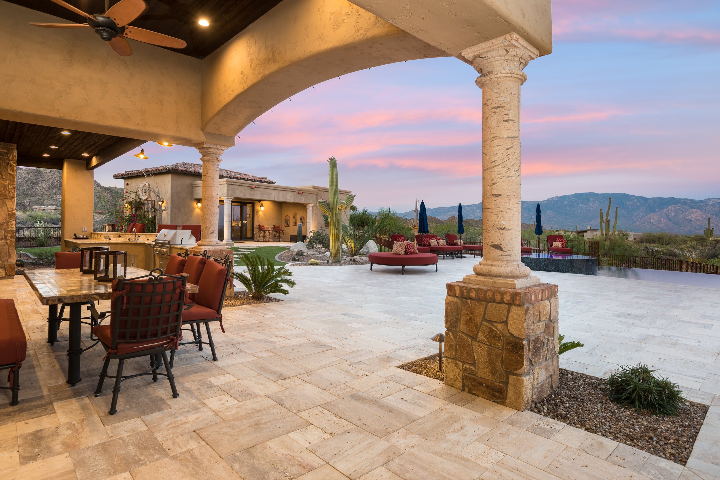 Luxury outdoor patio with seating and ceiling fan, stone pillars, desert landscape, mountain view, and modern outdoor kitchen at sunset.
