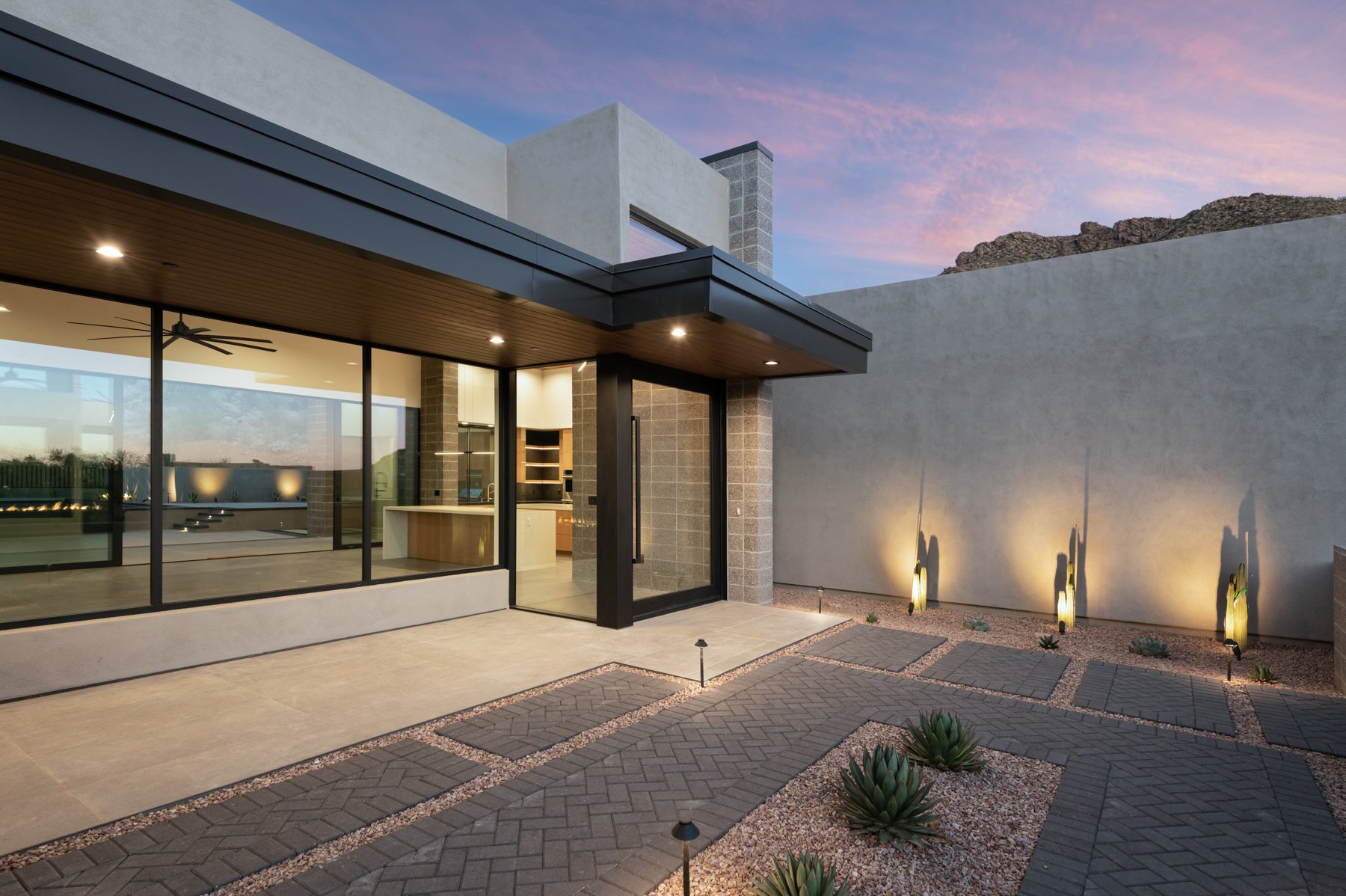 Modern house exterior with large windows, patio with decorative stone and plants, outdoor lighting, and a mountain in the background.